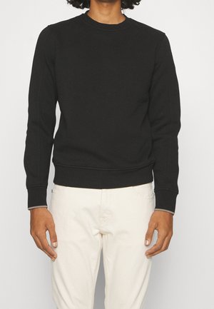 Persoon die een effen zwart sweatshirt met lange mouwen en een witte broek draagt, staand met ontspannen armen langs het lichaam tegen een lichte achtergrond.