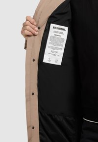 Mână ținând o jachetă bej deschisă, dezvăluind interiorul negru cu o etichetă de marcă rectangulară albă și un buzunar cu fermoar în interior.
