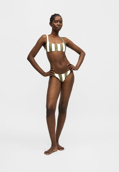 Γυναίκα όρθια ξυπόλητη με τα χέρια στη μέση, φοράει πράσινο και λευκό μπικίνι με κάθετες ρίγες, σε απλό ανοιχτό φόντο.