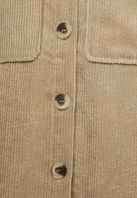 Beige kord skjorta med synlig ribbad textur, som har två bröstfickor och fyra runda knappar i en ljus, glänsande finish.