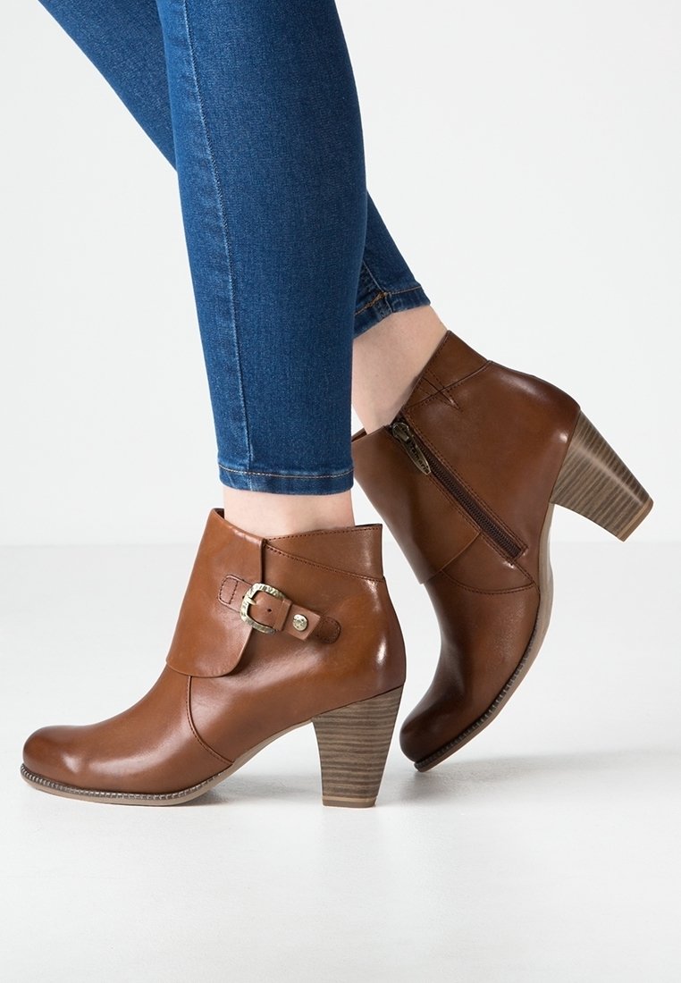Bottes courtes en cuir marron avec talons empilés, fermeture éclair latérale et détail de boucle, portées avec un jean bleu ajusté sur fond uni.