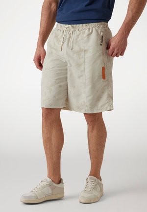 Beige shorts dengan pola halus, pinggang elastis, tali serut, dan saku zip samping dengan detail logo oranye. Desain kasual dan ringan.