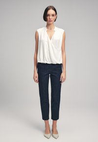 Blusa bianca senza maniche con scollatura a V profonda e orlo arricciato. Abbinata a jeans slim-fit blu scuro e décolleté a punta di colore chiaro.