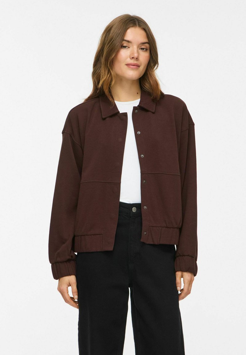 Jeune femme aux cheveux mi-longs portant une veste marron foncé boutonnée sur une chemise blanche et un pantalon noir, debout devant un fond uni.