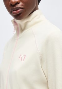 Gros plan d'une personne portant une veste crème zippée avec un col haut, une fermeture éclair rose et un logo sur la poitrine.