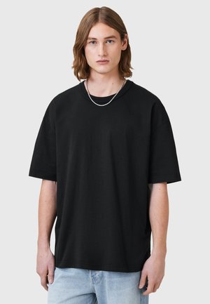 Black oversized cotton t-shirt with short sleeves, crew neckline, and a smooth texture, worn with a silver chain necklace.