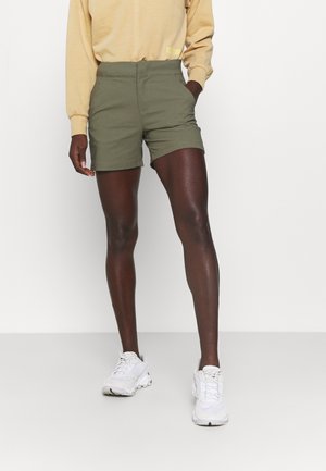 Personne portant un short vert olive, un haut à manches longues beige et des baskets blanches, debout devant un fond uni.