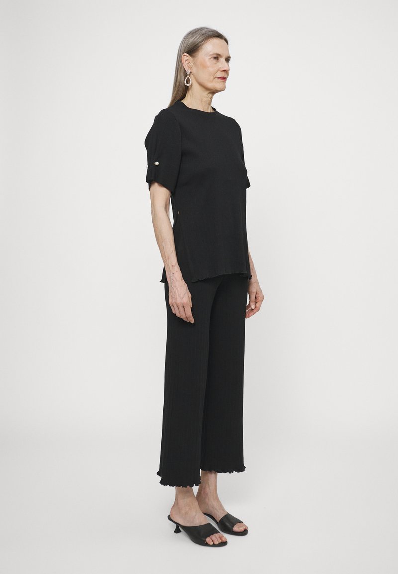 Black ribbed top with short sleeves and side slits, paired with matching cropped pants featuring scalloped hems. Black open-toe heels.