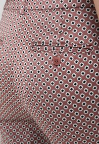 Pantaloni marroni con motivo a esagoni in nero e bianco. Presentano una tasca posteriore con dettaglio a bottone. Tessuto dalla texture liscia.