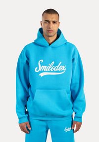 Sudadera turquesa con un gran logo blanco de "Smilodox", bolsillo frontal y capucha con cordón. Tejido suave y corte relajado, combinada con pantalones a juego.
