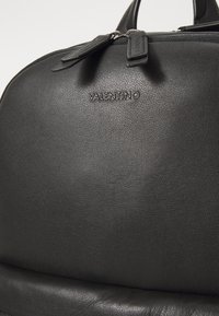 Zwarte leren rugzak met een gladde textuur, voorzien van een ritsafsluiting en een geperst "Valentino" logo aan de voorkant. Ronde vorm en stevige handvatten.
