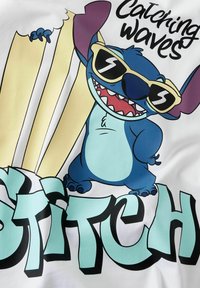 Maglietta bianca con un personaggio blu in stile cartone animato che indossa occhiali da sole, circondato da tavole da surf, con testo in grassetto che dice "Catching Waves" e "Stitch."