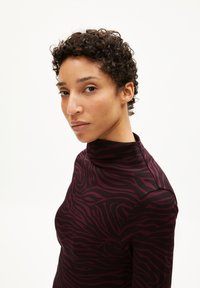 Frau mit kurzen, lockigen Haaren, die einen dunkelvioletten und schwarzen, zebra-gestreiften Rollkragenpullover trägt und leicht zur Seite schaut vor einem weißen Hintergrund.
