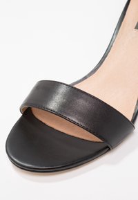 Gros plan de la partie avant d'une sandale en cuir noir à bout ouvert avec une seule large bride et une semelle intérieure beige.