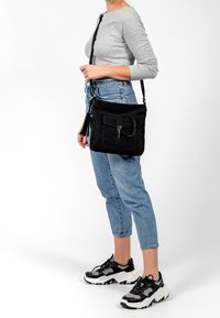 Schwarze Wildleder-Umhängetasche mit einer vorderen Klappentasche, silberner Hardware und Quasten-Detail. Kompakte Form, ideal für den legeren Alltag oder Reisen.