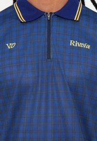 Blå og marinefarvet ternet polo shirt med lynlås krave. Har gule striber på kraven og broderet tekst "Rivoiă" og "VØ."