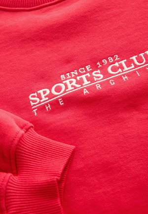 Punainen collegepaita, jossa on resorit hihansuissa ja kauluksessa. Etuosassa valkoisin kirjaisin tekstein "SPORTS CLUB" ja "SINCE 1982". Pehmeä pinta.