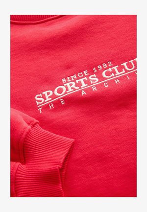 Rdeč pulover s patentnimi manšetami in ovratnikom, na sprednji strani z belim vezenim napisom "SPORTS CLUB" in "SINCE 1982". Mehka tekstura.