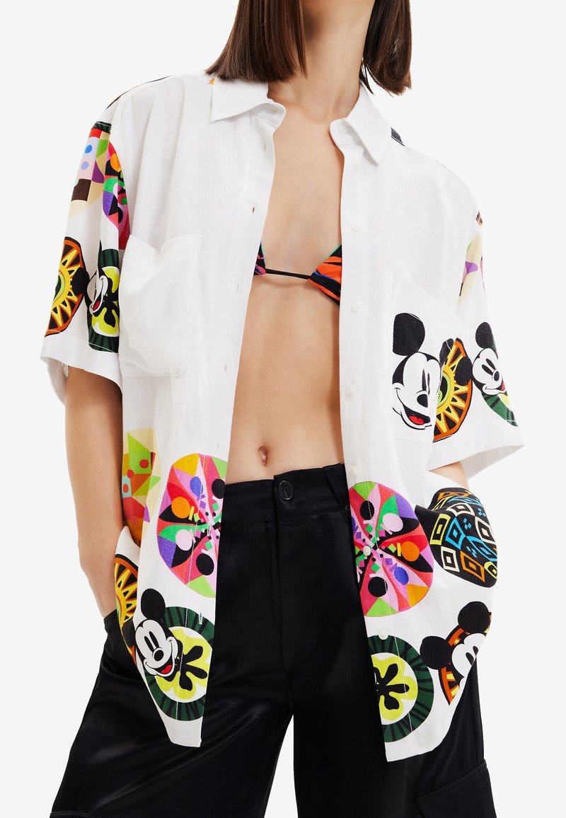 Personne portant une chemise blanche ouverte avec des imprimés colorés de Mickey Mouse sur un haut de bikini noir et orange et un pantalon noir.