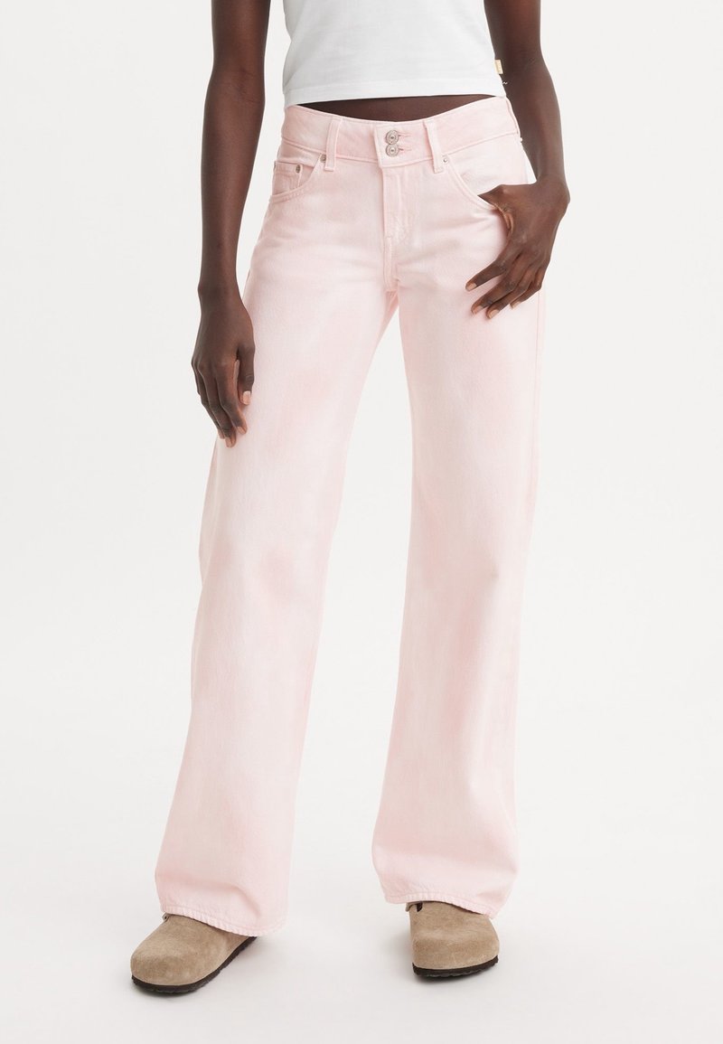 Personne portant un jean large rose clair, un top court blanc et des mocassins en daim beige, se tenant avec une main dans la poche.
