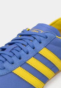 Blå joggesko med gule striper, glatt tekstur, rund tå og snøring. Detaljer inkluderer Adidas-logo og sømmer.