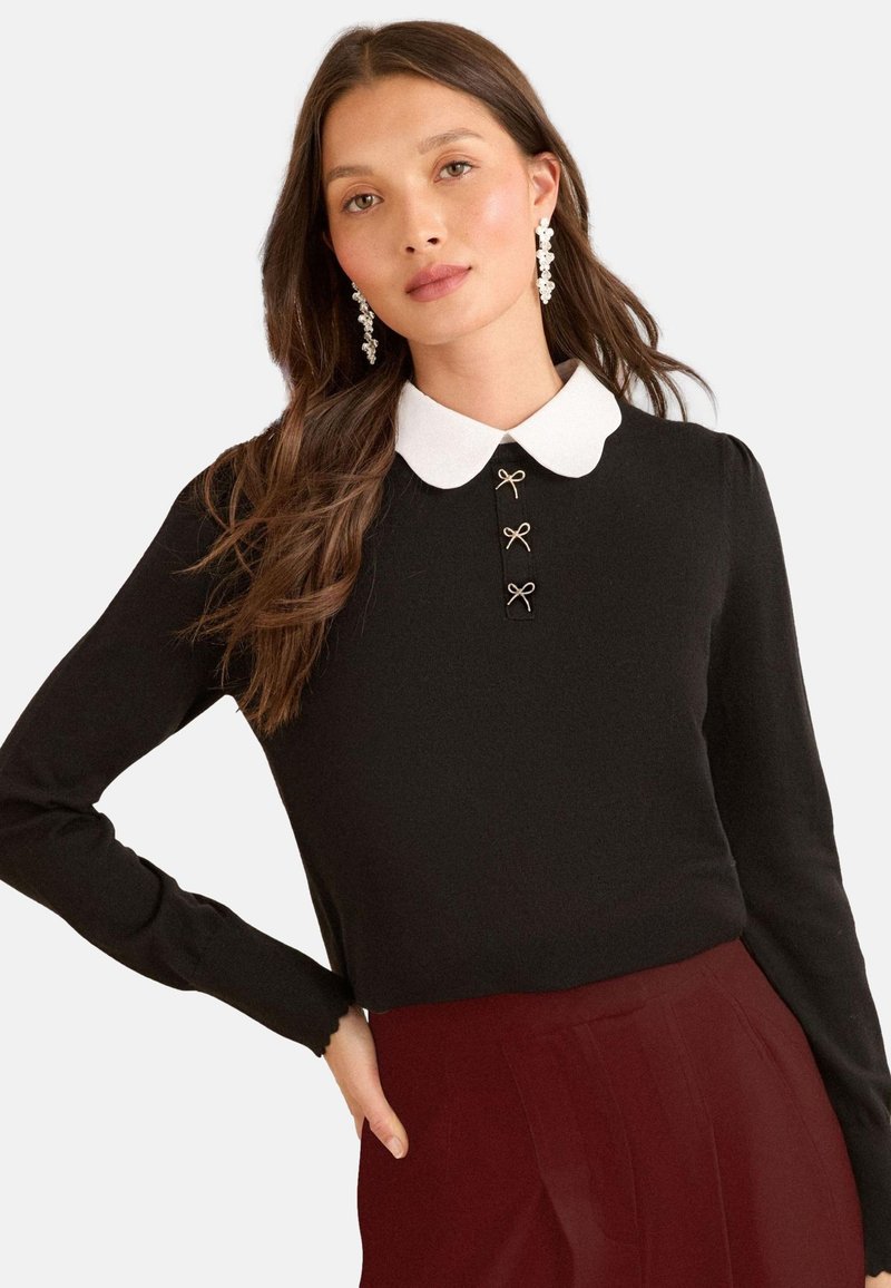 Woman with long brown hair wearing black long-sleeve top with white rounded collar and three small bow details, paired with burgundy pants.