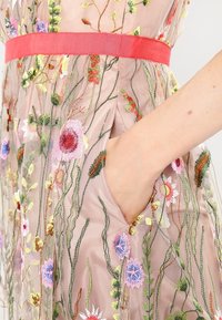 Genomskinlig, broderad klänning med blommönster i livliga färger. Har en åtdragen röd midja och sidofickor. Strukturerad tyg.