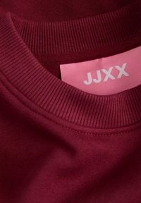 Collo di felpa in cotone bordeaux con texture a coste, dotato di un'etichetta rosa su cui è scritto "JJXX" in lettere bianche.