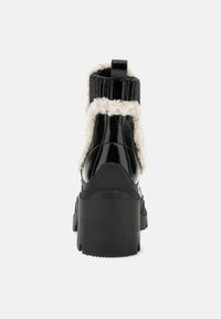 Botas de tobillo de charol negro con un cuello blanco esponjoso, parte superior de punto acanalado y suela texturizada. En la parte posterior se muestra la marca "GUESS". Tacón grueso.