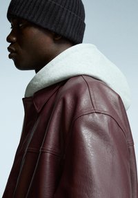 Veste en cuir bourgogne avec un col classique, portée sur un sweat à capuche gris clair. L'arrière-plan est un dégradé neutre.