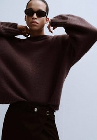 Hnědý vlněný svetr s opadanými rameny a žebrovanými manžetami, doplněný černými vysokému pasu kalhotami s akcenty stříbrných knoflíků.