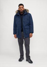 Parka azul marino con capucha de piel sintética, dos bolsillos delanteros y cierre con cremallera, llevada sobre una camisa negra y pantalones grises. Zapatillas negras completan el look.