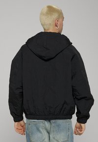 Giacca trapuntata nera con cappuccio, dotata di polsini elastici e orlo elasticizzato. Il tessuto presenta un motivo strutturato, indossata sopra jeans a lavaggio chiaro.