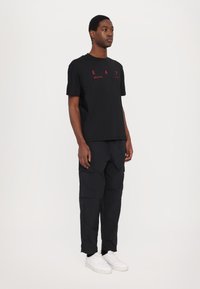 Black T-shirt with red logo detail, paired with black cargo pants and white sneakers. Features a crew neckline and relaxed fit.