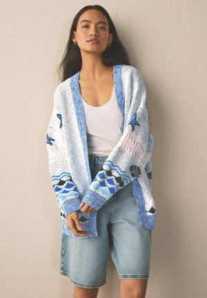 Junge Frau, die eine hellblaue gemusterte Strickjacke über einem weißen Tanktop und Jeansshorts trägt, vor einem beigen Hintergrund stehend.