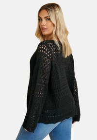Schwarzer gehäkelter Pullover mit einem offenen Strickmuster, oversize Passform und weiten Ärmeln, kombiniert mit blauen Jeans. Strukturierte Designdetails überall.