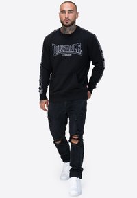 Czarna bluza z logo "Lonsdale London", długie rękawy z wzorzystymi akcentami, w połączeniu z postarzanymi czarnymi dżinsami i białymi trampkami.