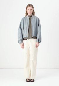 Giubbotto bomber in pelle grigia chiara con zip frontale, colletto a coste e vestibilità ampia, abbinato a pantaloni a gamba larga color crema e sandali neri.