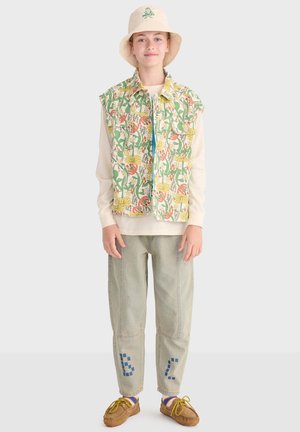 Barn iført en vest med blomsterprint, cremefarvet langærmet skjorte, lyse denimbukser med blå lapper, en beige bøllehat og brune mokkasiner.