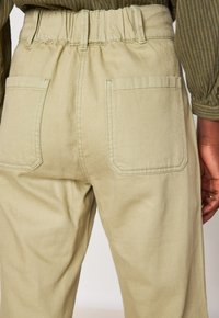 Beige broek met een elastische tailleband en achterzakken, gedragen met een bruine geribbelde long-sleeve top aan de rechterkant.