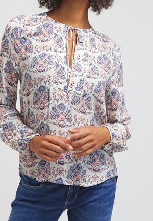 Vrouw draagt een blouse met lange mouwen met een blauw en roze bloemenpatroon, een keyhole-halslijn met strikjes, gecombineerd met een blauwe spijkerbroek.