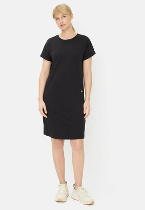 Blondynka stojąca, ubrana w czarną sukienkę z krótkim rękawem sięgającą do kolan oraz białe trampki na białym tle.