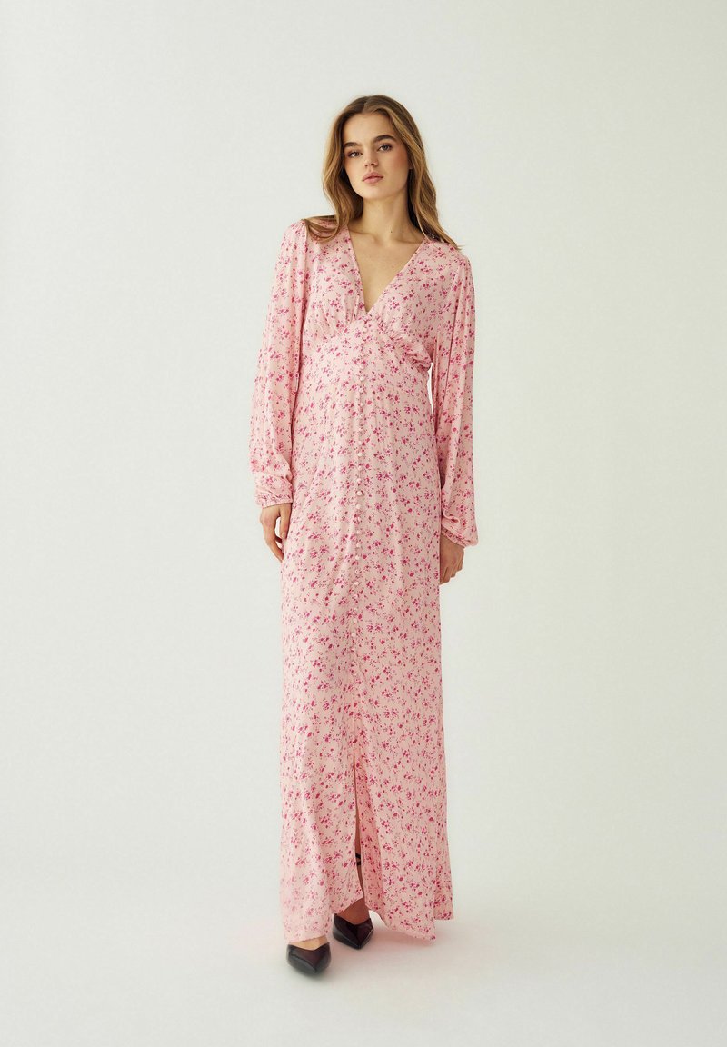 Naine seisab seljas pikk roosa lillemustriga kleit pika varruka ja mustade teravate kingadega ühtlase heleda tausta ees.