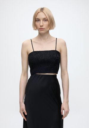 Tânără cu păr blond scurt purtând un top scurt negru cu flori și o fustă în talie asortată, pe un fundal simplu și deschis la culoare.