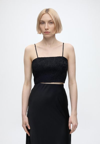 Jeune femme aux cheveux blonds courts portant un haut court noir à motifs floraux et une jupe taille haute assortie sur un fond clair uni.