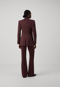 Traje burdeos a medida con americana ajustada y pantalones acampanados, hecho de un tejido texturizado, con una espalda estructurada y líneas elegantes.