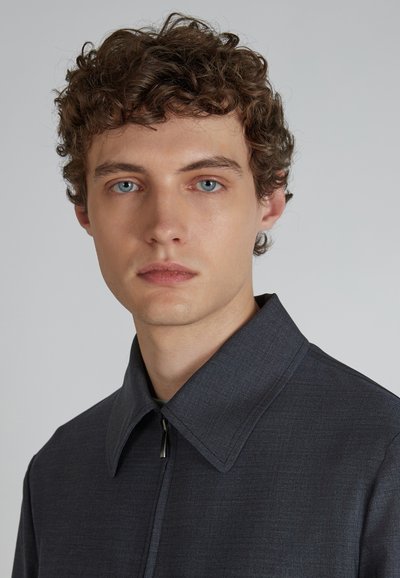 Ciemnoszara kurtka z kołnierzykiem, wykonana z teksturowanej tkaniny, z zamkiem błyskawicznym z przodu i dopasowanym krojem. Zbliżenie na górną część ciała i dekolt.