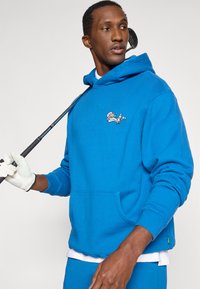 Μπλε φούτερ από βαμβάκι, με μπροστινή τσέπη και κεντημένο γραφικό ενός χαρακτήρα που καπνίζει πίπα. Φοριέται με γάντι γκολφ.