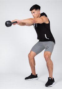 Black sleeveless hoodie, gray athletic shorts with logo, performing a kettlebell exercise. Black athletic shoes, neutral background.