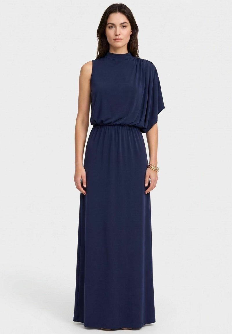 Femme portant une robe longue sans manches bleu marine avec un col haut et un tissu drapé asymétrique sur une épaule, debout, les bras détendus.
